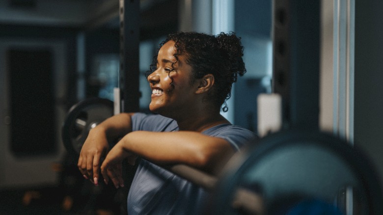 A person in a gym leans on a barbell, smiling, with a blurred background indicating equipment and dim lighting in the scene.