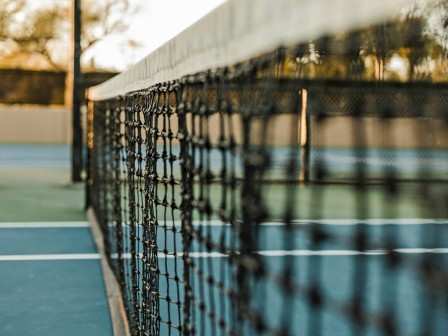 A close-up of a tennis court net with blurred background, showing the court's surface and surrounding fence or barrier in the distance.