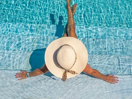 A person in a wide-brimmed hat relaxes in a shimmering blue pool, arms outstretched, enjoying a sunny day in the water.