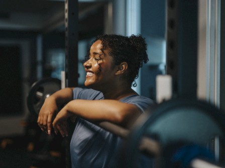 A person is smiling and leaning on a barbell in a gym setting, with a relaxed and cheerful expression.