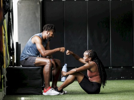 Two people in workout attire are sitting in a gym, exchanging a fist bump, one holding a water bottle on green turf.