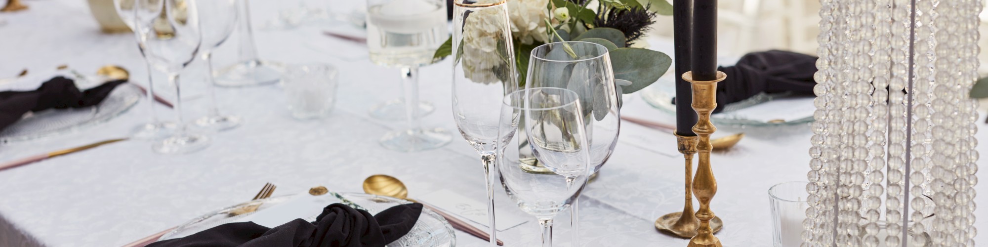 A elegantly set table features black napkins, glassware, gold cutlery, floral centerpieces, and black candles, creating a chic atmosphere.