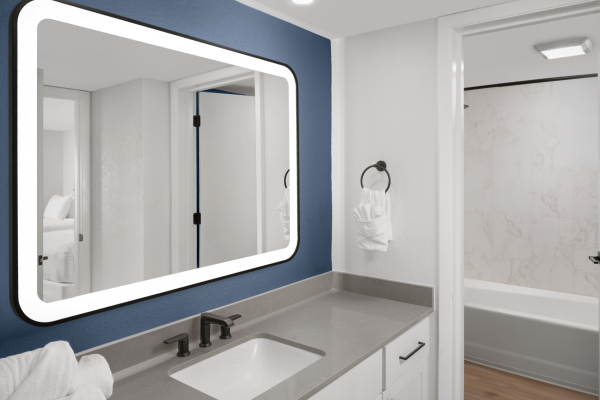 A modern bathroom with a large lit mirror, sink, and a glimpse of a bathtub in the adjacent room.