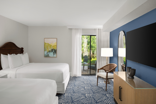 The image shows a hotel room with two beds, a TV, and a balcony view. The decor is modern with blue and white tones.