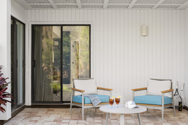 A patio with two chairs, a small table holding drinks, a cap, and a book, set against a white wall with a glass sliding door.