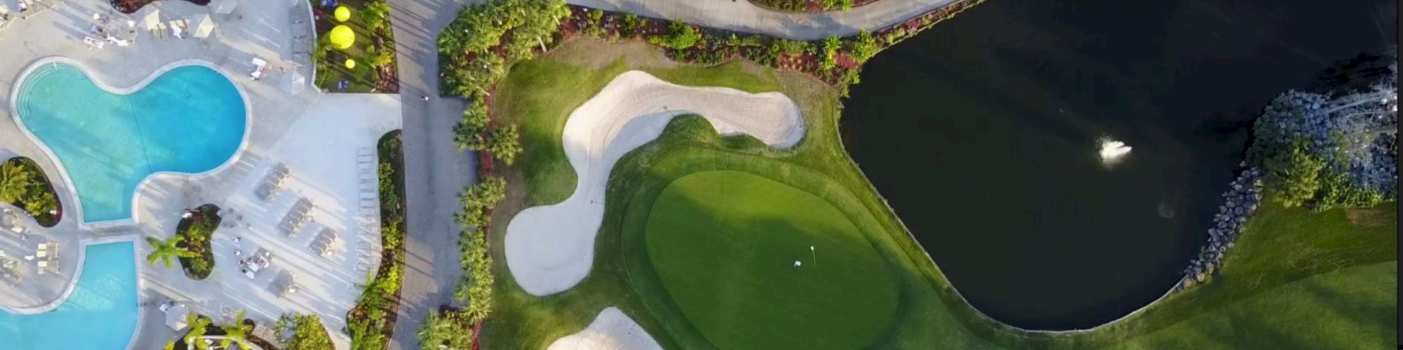 The aerial view shows a pool area, a road, a golf course, and a small lake with trees and pathways surrounding them.
