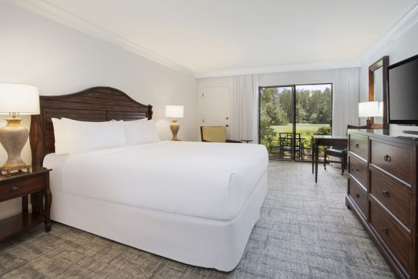 A neat hotel room with a bed, lamps, dresser, TV, desk, chair, and a view outside the window.