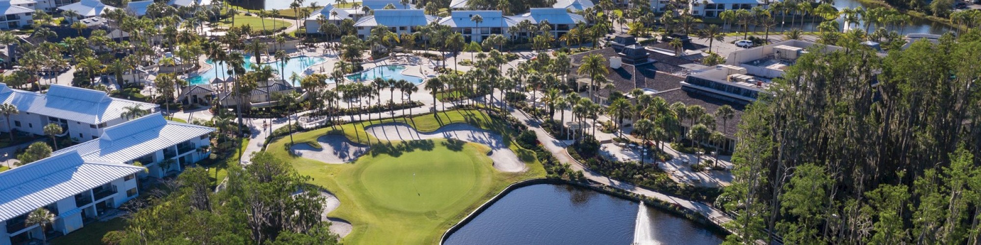 Aerial view of a resort with pools, green lawns, and a picturesque lake with a small sailboat, surrounded by trees and buildings.