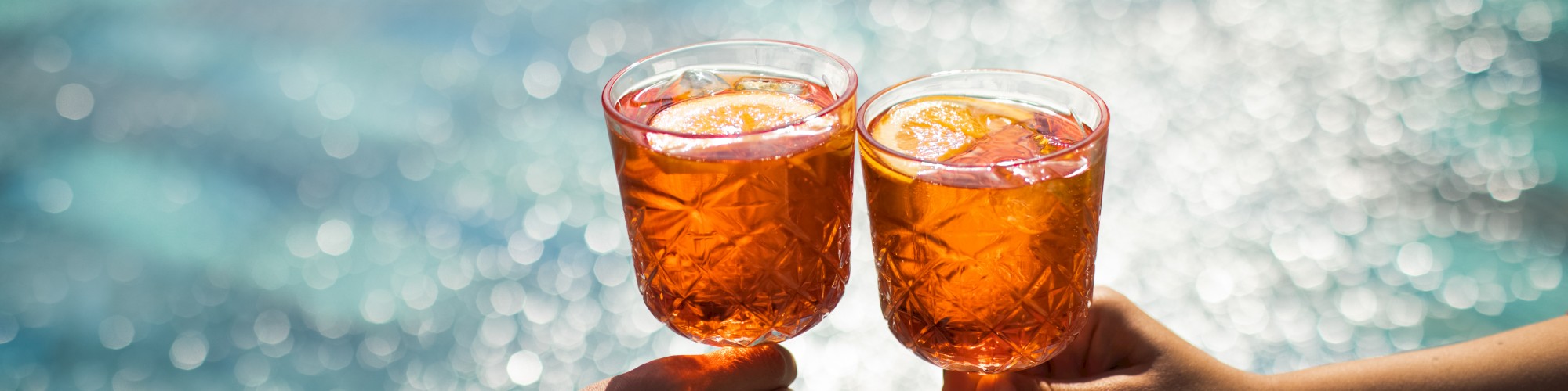 Two hands holding glasses with orange drinks, clinking them together in front of a sparkling pool background.