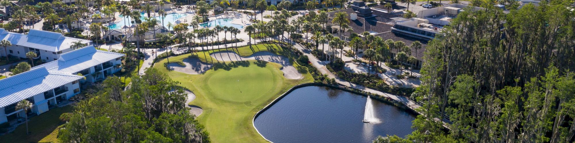Aerial view of a golf course with a pond beside resort-style buildings and pools