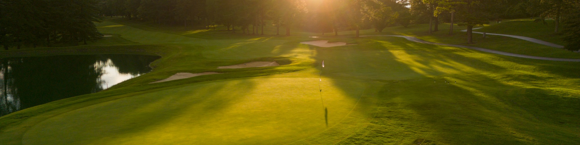 A golf course at sunset, with lush green fairways, sand traps, and a water hazard, surrounded by trees.