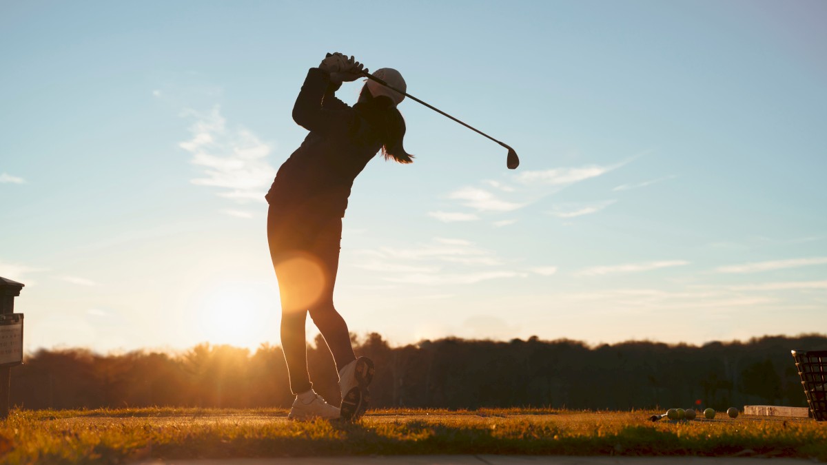 A person is playing golf, swinging a club at sunset on a grassy field with trees in the distance, under a clear sky.
