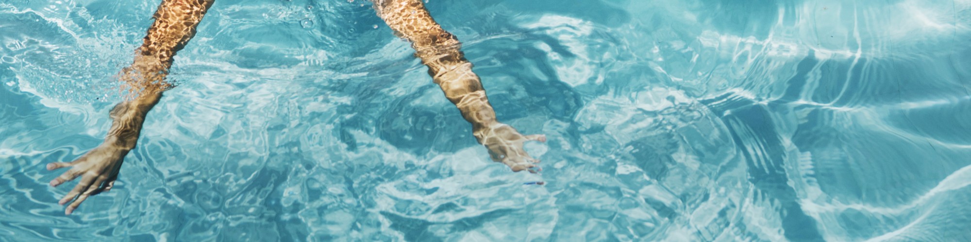 A pair of legs submerged in a swimming pool with clear blue water and ripples forming around them, under sunlight, creating a serene view.