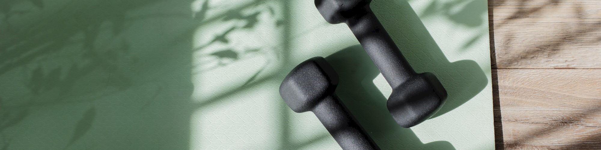 Two black dumbbells on a green yoga mat with shadows of leaves, creating a peaceful workout ambiance.