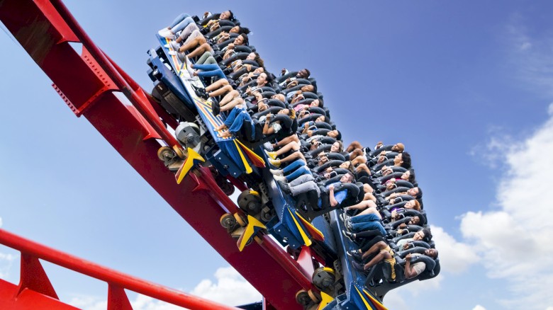 A roller coaster with people riding, faces blurred in motion, on a steep red track against a bright blue sky with scattered clouds.
