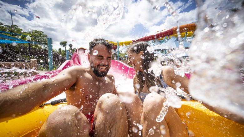Two people enjoy a water slide, splashing around on a bright, sunny day at a water park.