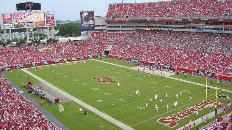 A packed stadium with a football game in progress, featuring red and white-clad players. There is a large scoreboard and many spectators.