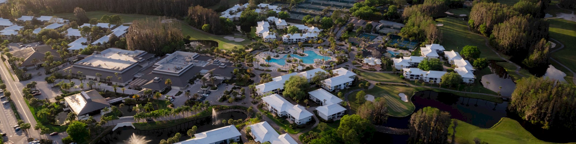 An aerial view of a resort complex surrounded by greenery, with buildings, a pool, and nearby roads under a partly cloudy sky.