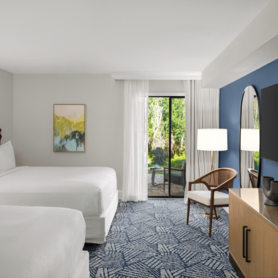 A modern hotel room features two beds, a flat-screen TV, a wooden chair, a lamp, a cabinet, artwork, and a view to an outdoor area.