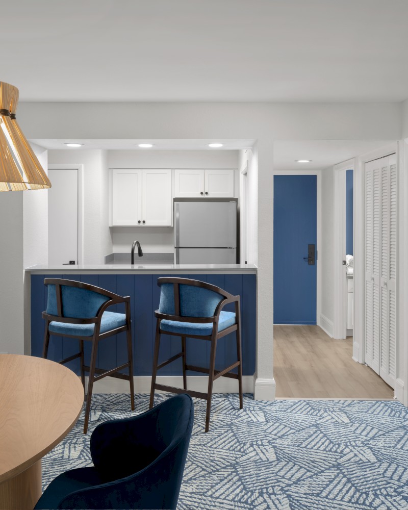 A modern kitchen and dining area with a wooden table, wicker light fixture, blue chairs, and a partitioned kitchen in view.