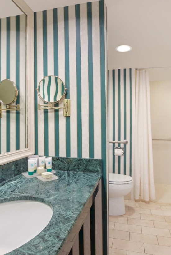 A bathroom with striped wallpaper, mirror, sink, and a visible shower area.