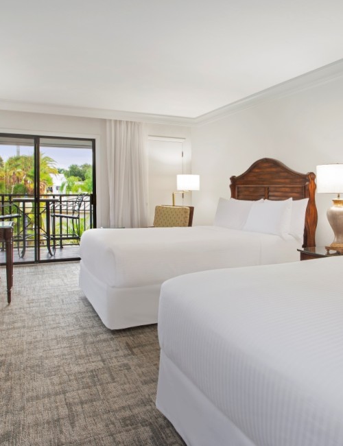 A clean, well-lit hotel room with two beds, a TV, desk, and a balcony view.