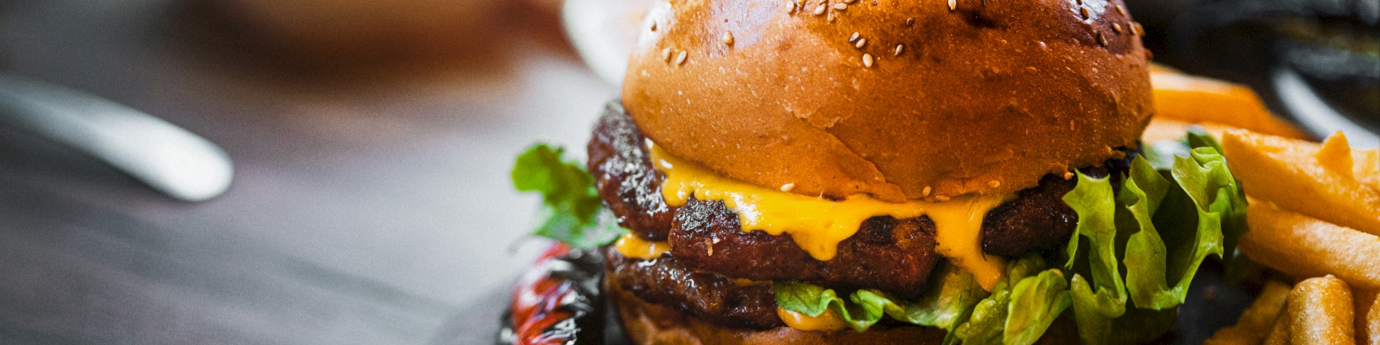 A cheeseburger with lettuce and sauce sits on a wooden board, accompanied by fries and a pepper, with sauces in the background.