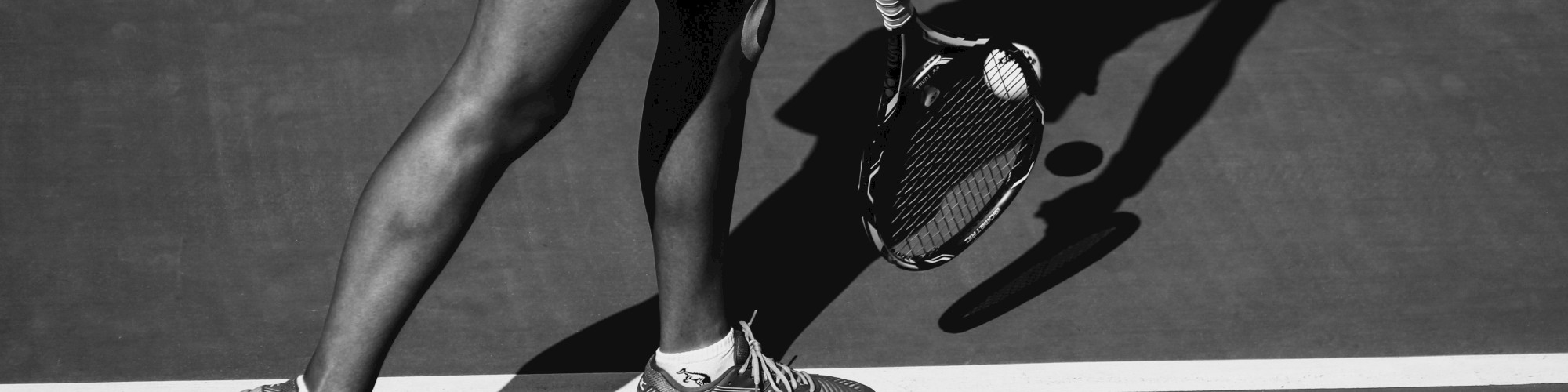 A person playing tennis, captured in black and white, mid-action with the shadow prominently visible on the court surface.