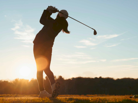 A person is playing golf at sunset, swinging a club on a grassy course with a clear sky and trees in the background.
