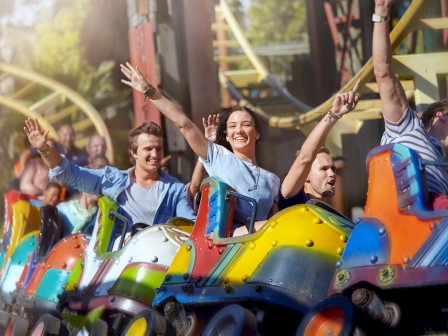 People enjoying a colorful roller coaster ride, smiling and raising their hands, with tracks and greenery visible in the background.