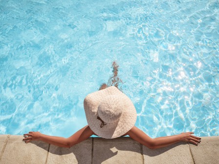 A person wearing a sun hat relaxes at the edge of a swimming pool with clear blue water.