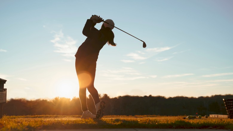 A person is silhouetted swinging a golf club against a sunset backdrop on a golf course, surrounded by trees and open landscape.