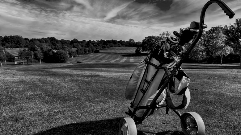 A monochrome image of a golf bag on a trolley on a lush golf course under a dramatic sky with streaky clouds.