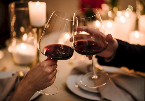 Two people clinking red wine glasses over a candlelit dinner table set with plates and napkins.