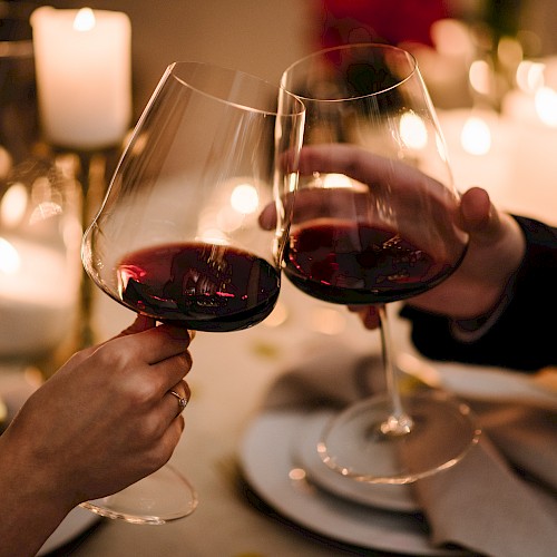 Two people clinking red wine glasses over a candlelit dinner table set with plates and napkins.