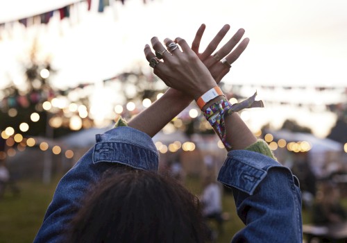 A person with raised arms at an outdoor festival, wearing bracelets and rings, with lights and flags in the background, during the evening.