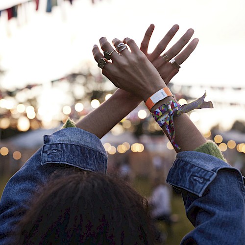 A person with raised arms at an outdoor festival, wearing bracelets and rings, with lights and flags in the background, during the evening.