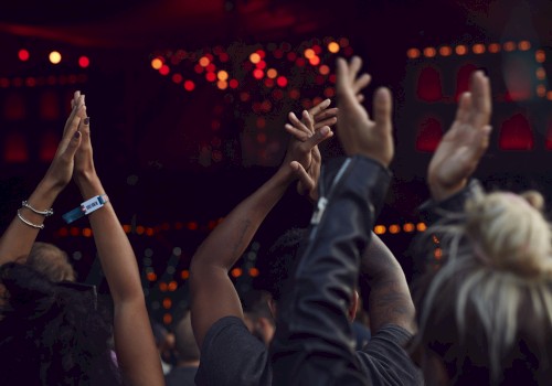 People raising their hands in the air at a concert or event, with vibrant red and orange lights in the background.