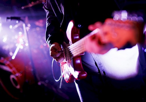 A person playing an electric guitar on stage, with a drum set and purple lighting in the background, creating a vibrant concert scene.