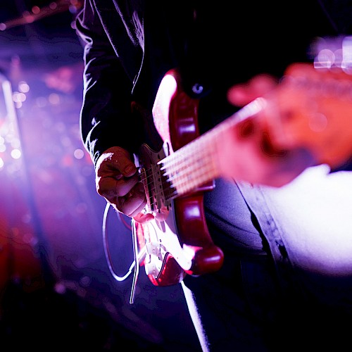 A person playing an electric guitar on stage, with a drum set and purple lighting in the background, creating a vibrant concert scene.