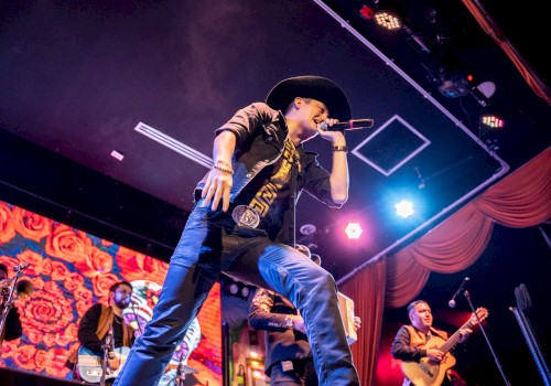 A performer in a cowboy hat sings on stage with a band, vibrant visuals behind them, under colorful lights creating an energetic atmosphere.