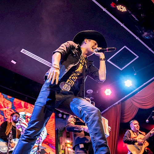 A performer in a cowboy hat sings on stage with a band, vibrant visuals behind them, under colorful lights creating an energetic atmosphere.