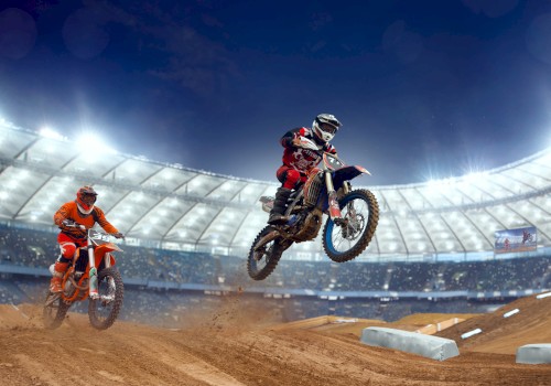 The image shows two motocross riders racing on a dirt track inside a brightly lit stadium, with one rider airborne.