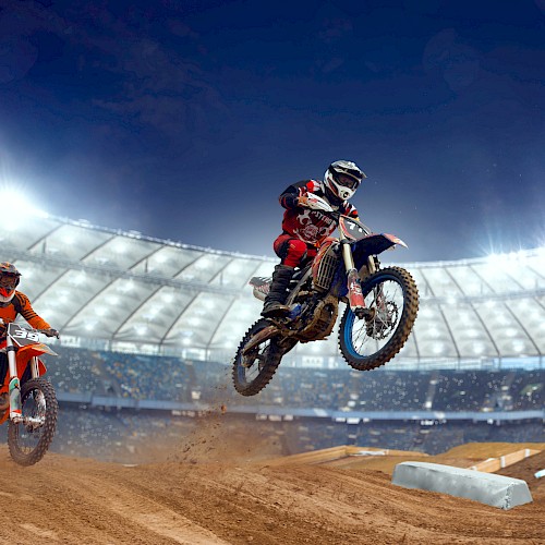 The image shows two motocross riders racing on a dirt track inside a brightly lit stadium, with one rider airborne.