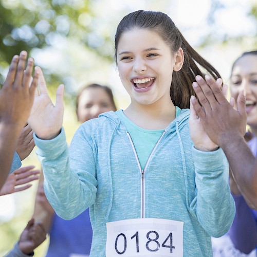 A girl with the number 0184 on her clothes is high-fiving people. She appears happy and is surrounded by others outdoors.