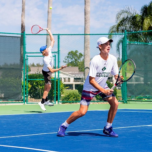 Two people are playing tennis on an outdoor court. One is serving, the other is ready to receive the ball.