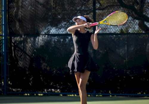 A person is playing tennis, wearing a black outfit and hat, holding a tennis racquet against a dark background on a sunny day.