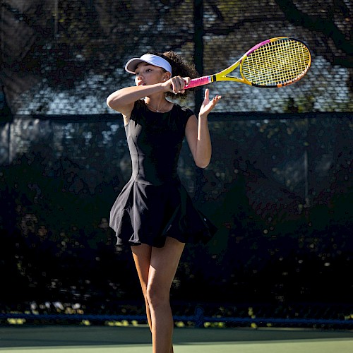 A person is playing tennis, wearing a black outfit and hat, holding a tennis racquet against a dark background on a sunny day.