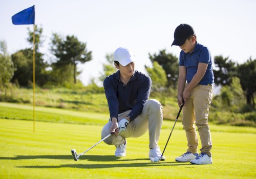 Two people on a golf course; one kneels, instructing the other on putting. Blue flag in the background, sunny day.
