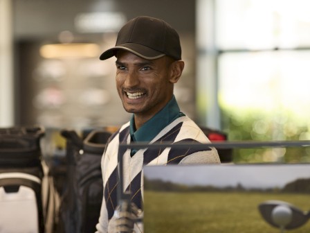 A person wearing a cap and argyle sweater is smiling indoors, near golf-related equipment and a picture of a golf course.